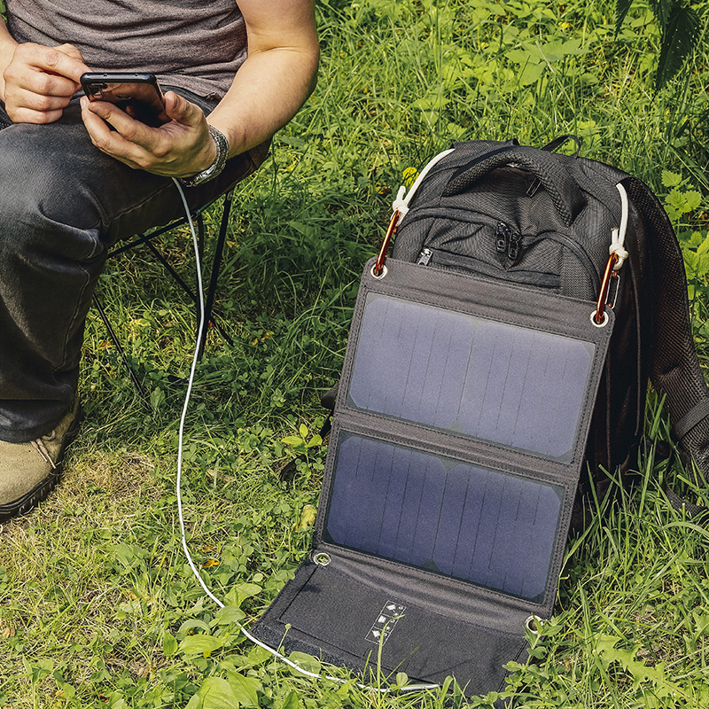 A camper is sitting on a camping chair and looking at smartphone that is charging from a solar panel next to a backpack on sunny summer day