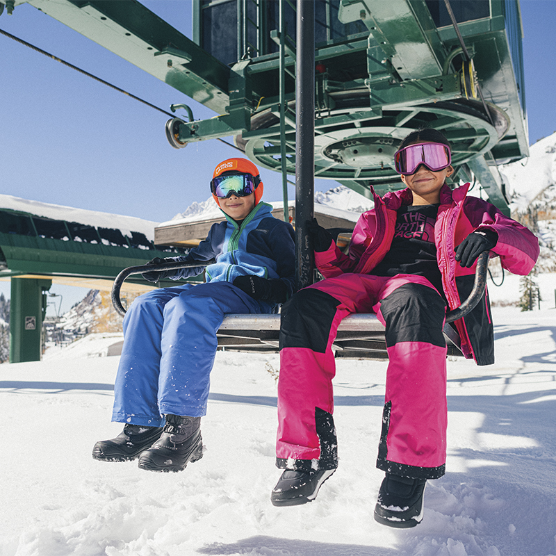 Two children sit on a ski lift in their ski and snowboard gear